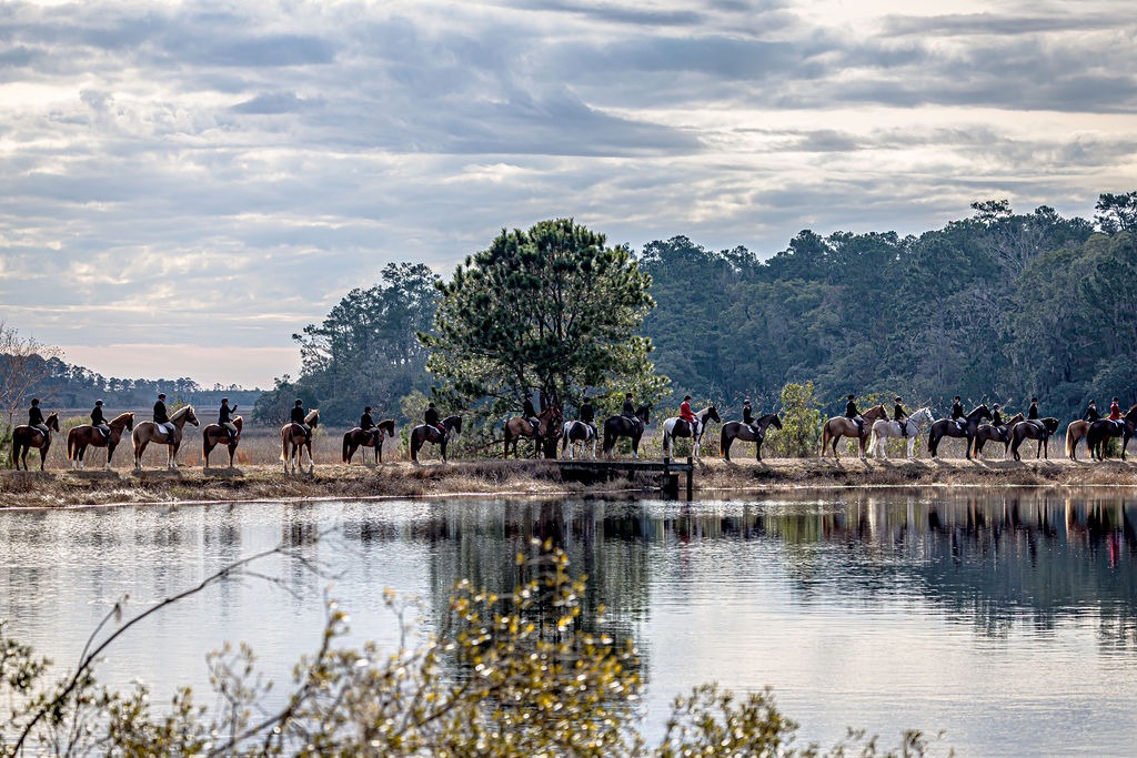 A long line of mounted riders beside a pond, reflected in the water beneath a cloudy sky.
